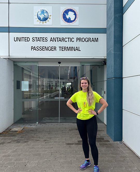 Photo of Lauren posing in front of building entrance with signage that says United States Antarctic Program Passenger Terminal